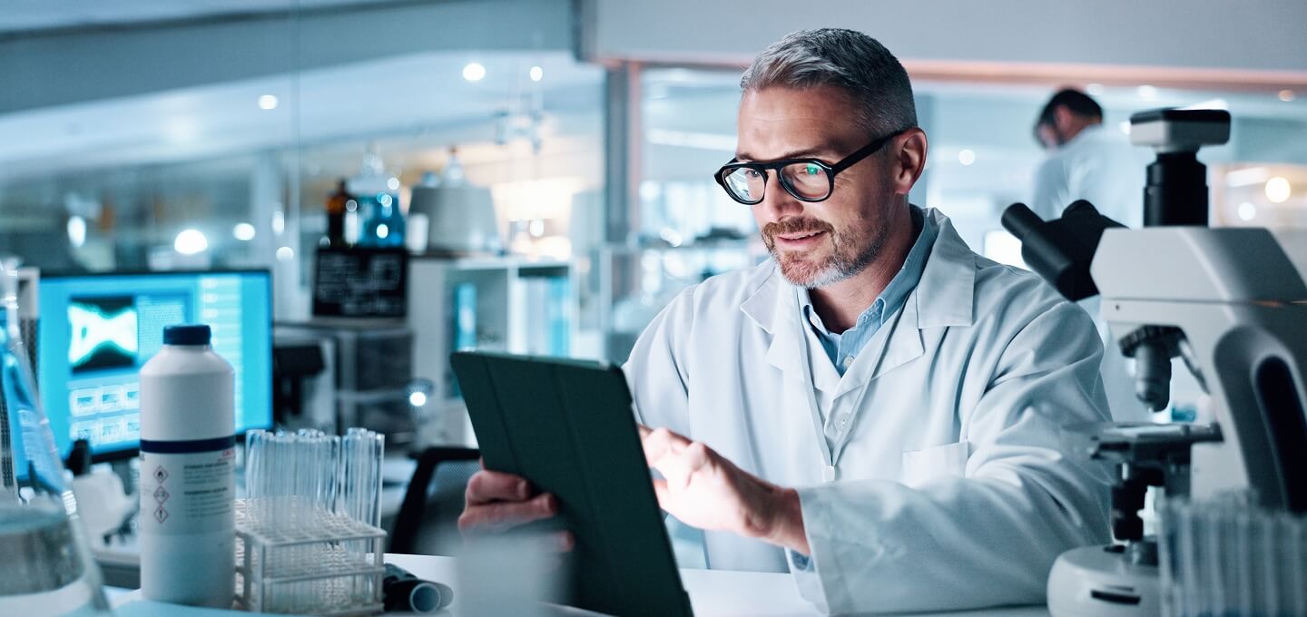 Scientist wearing glasses and a white lab coat reviews data on a tablet in a modern laboratory surrounded by microscopes, test tubes, and monitors.