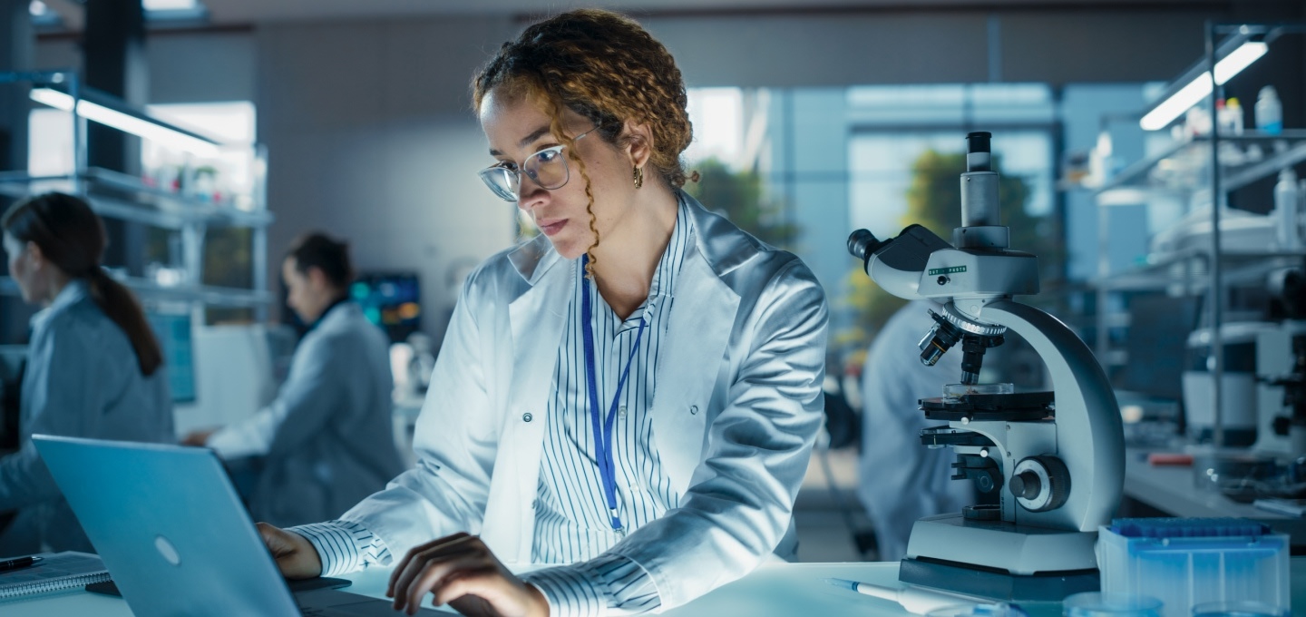A woman in a white lab coat using a laptop next to a microscope in a high-tech laboratory setting.