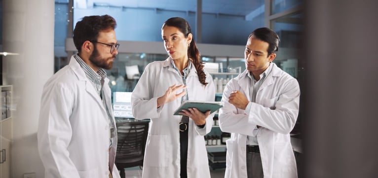 Three scientists in white lab coats discuss findings on a tablet during a collaborative meeting in a modern research laboratory.