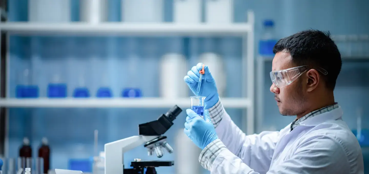 Male researcher in a pharmaceutical laboratory, wearing protective gloves, safety goggles, and a white lab coat, precisely adding liquid into a beaker using a dropper, with laboratory tools and equipment like a microscope and chemical-filled containers in the background.